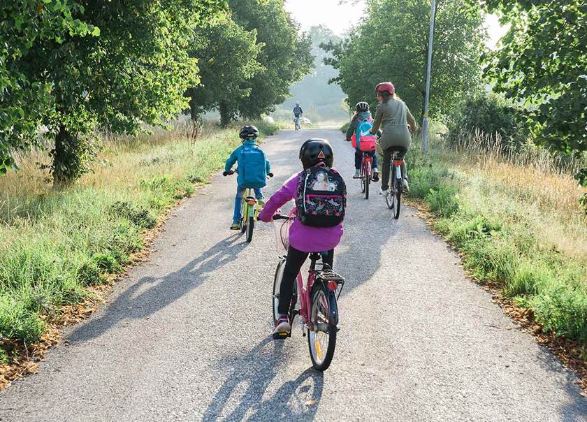 Cyklande barn och vuxen på grusväg omgiven av ängsmark och skog.