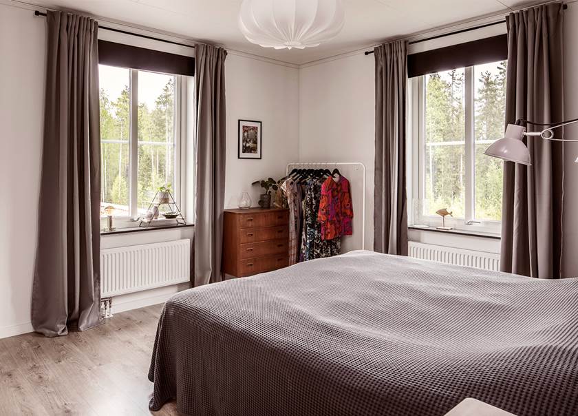 Sovrum med två fönster med grå gardiner dubbelsäng antik byrå klädhängare hemma hos familjen Engström