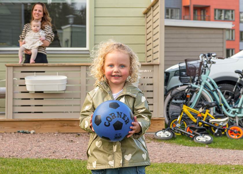 Flicka i blont lockigt hår och beige regnjacka med vita stjärnor står med blå fotboll i händerna.