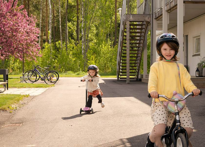En slicka på cykel och en flicka på sparkcykel framför radhus