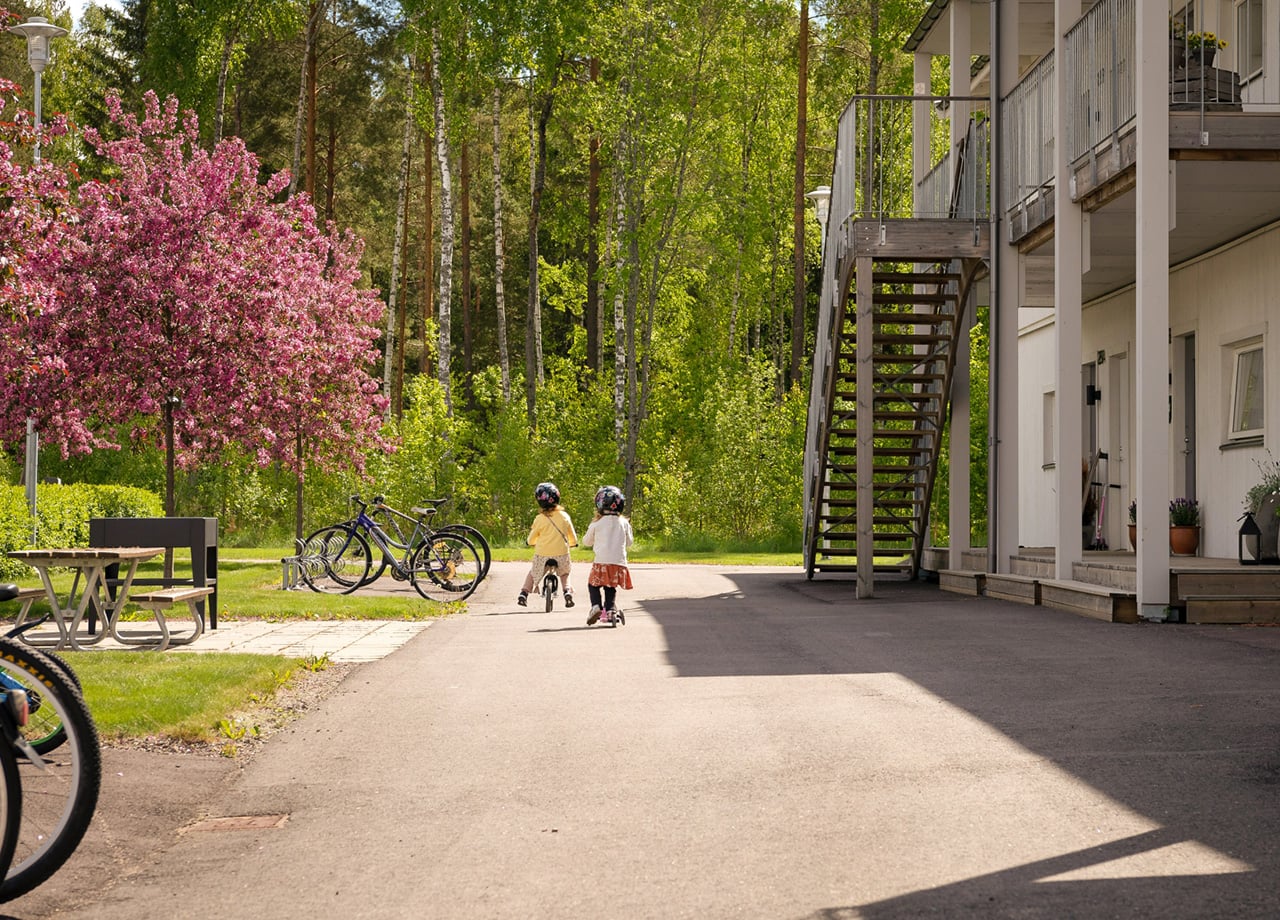 En flicka på cykel och en på kickbike på väg bort på gångväg intill ett lägenhetshus entrésida