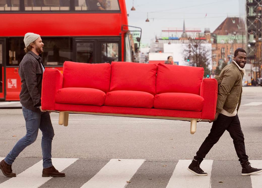 två yngre män bär på en röd soffa över ett övergångsställe mitt i stan