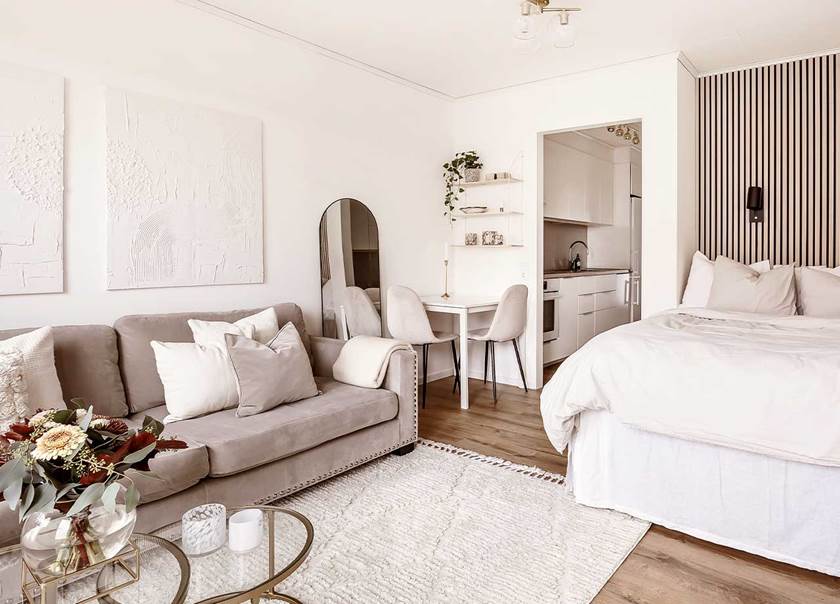 Vy av kök och rum i en etta med beige soffa, vit ryamatta, dubbelsäng med sänggavel av akustikpanel i ljust trä.