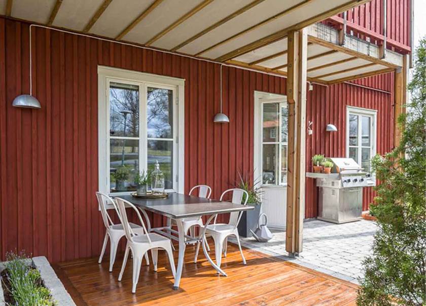 Terass med trä och sten, med vita stolar och matbord mot röd trävägg Terass med trä och sten, med vita stolar och matbord mot röd trävägg