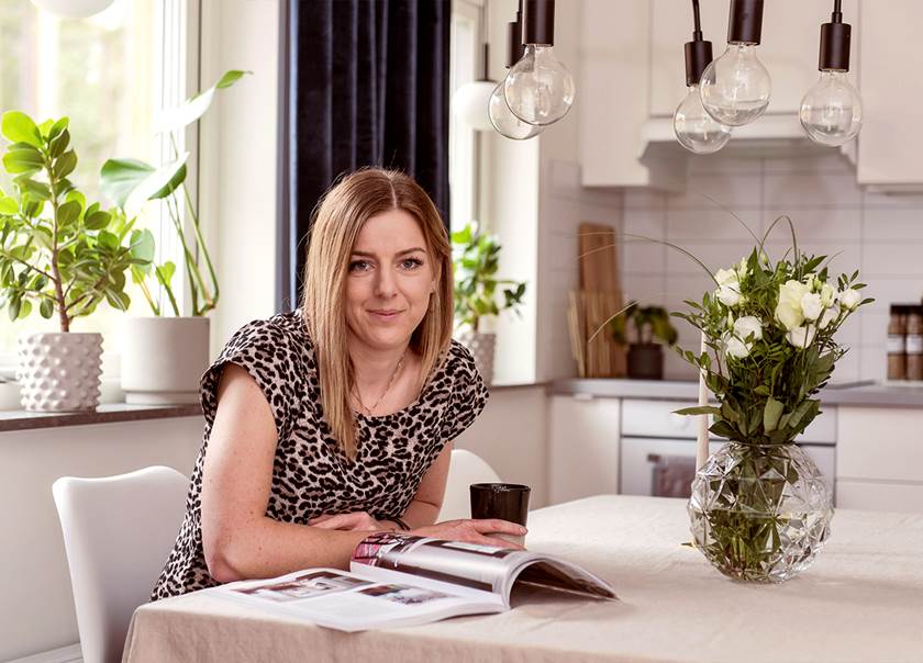 kvinna med mellanblont hår sitter och läser inredningstidning vid ett matbord med vit duk och vita rosor i en vas kvinna med mellanblont hår sitter och läser inredningstidning vid ett matbord med vit duk och vita rosor i en vas