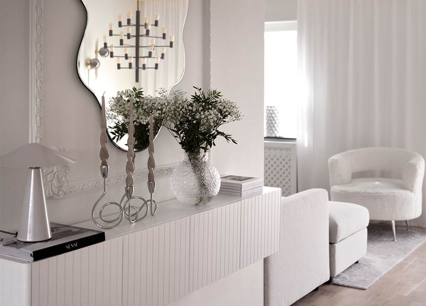 Lampa, cirkelformade silverljusstakar, vas med vita blommor på vägghängt vitt skåp. Ovanför hänger en spegel.