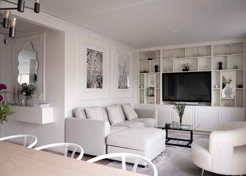 Vardagsrum med ljusgrå soffa och vit inbyggd bokhylla och stuckaturer på väggar och i taket.