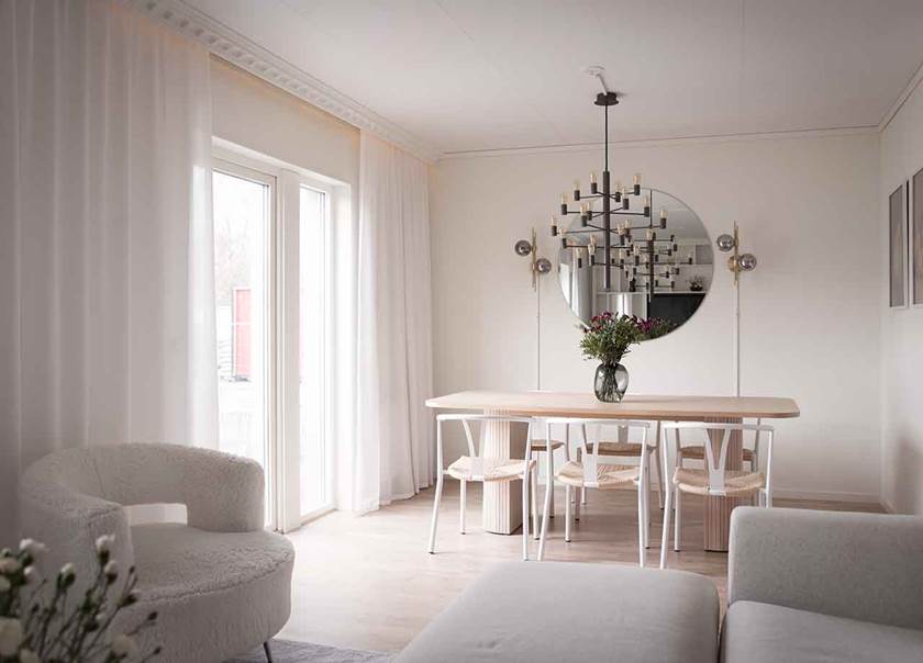 Ljusgrå soffa i förgrund och matbord i ljust trä med vita stolar runtom med flätad sits i rotting i vardagsrum.