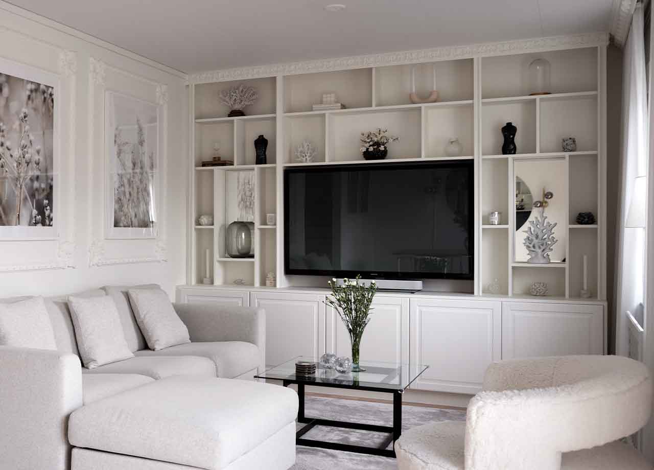 Vit bokhylla som räcker hela vägen upp till tak med vaser och stor TV i mitten, i vardagsrum med ljusgrå soffa.
