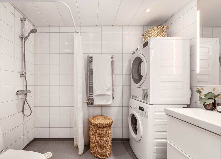 Badrum i vitt med toalett, tvättmaskin och torktumlare. I mitten står en tvättkorg av rotting.