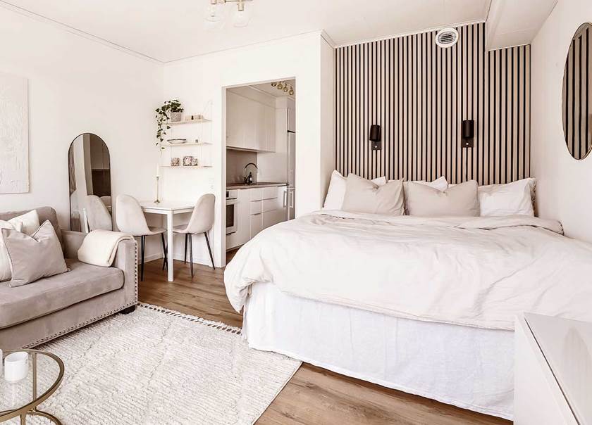 Vy av rum med beige soffa, matplats med två sammetsstolar och en dubbelsäng med akustikpanel i ljust trä som sänggavel.