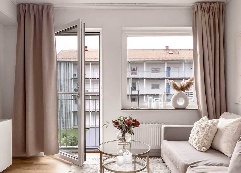Beige långa gardiner hänger på vardera sida av ett stort vardagsrumsfönster och en helglasad balkongdörr som står öppen ut mot en fransk balkong.