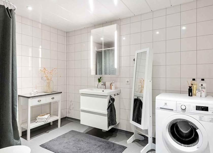Tvättmaskin och stor spegel på fötter i vitkaklat badrum med grå badrumsmatta, grå duschdraperi och vit förvaringsmöbel med grå marmorskiva.