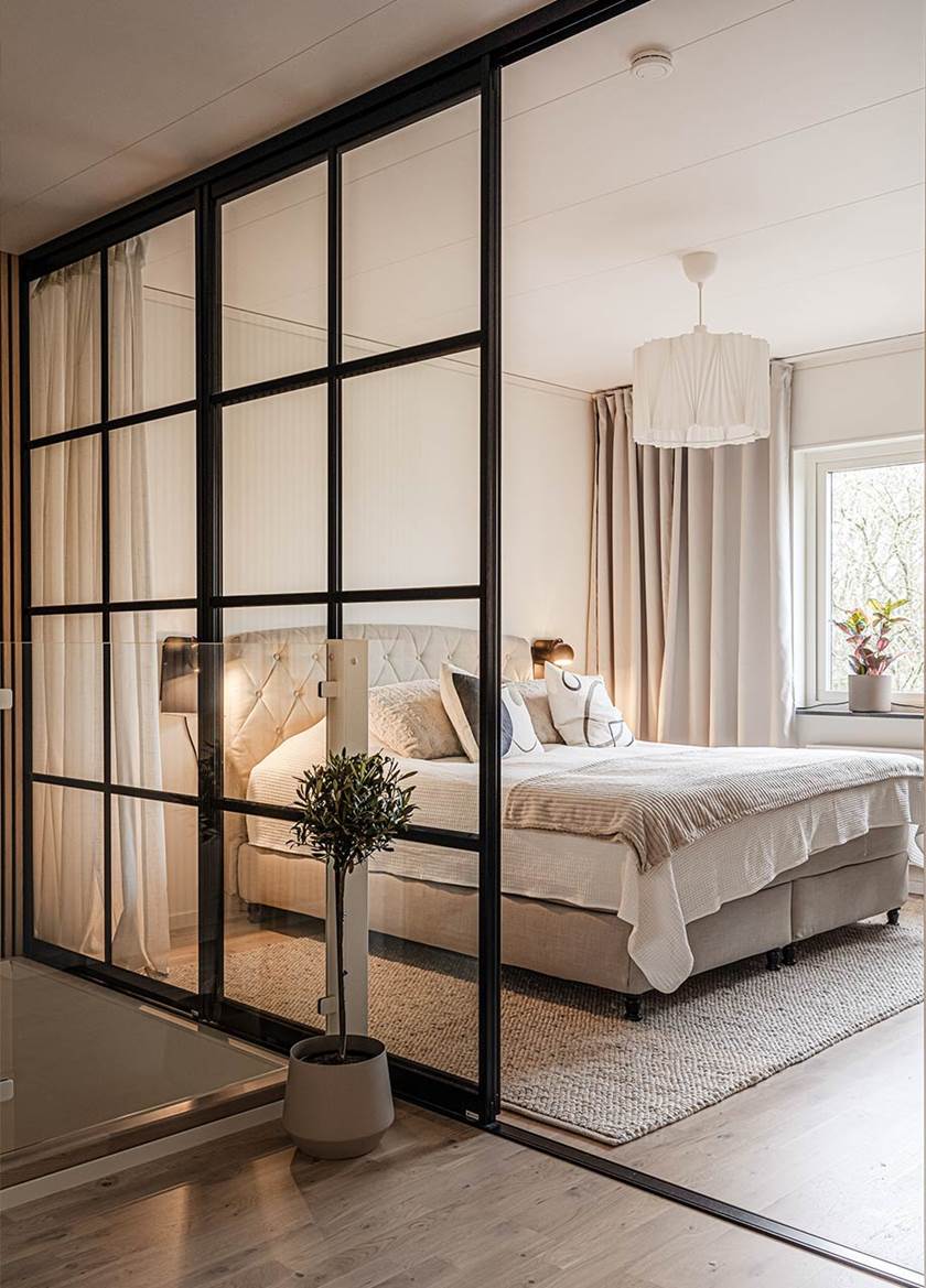 Vägg av glas och svarta stålramar intill ett sovrum med dubbelsäng.
