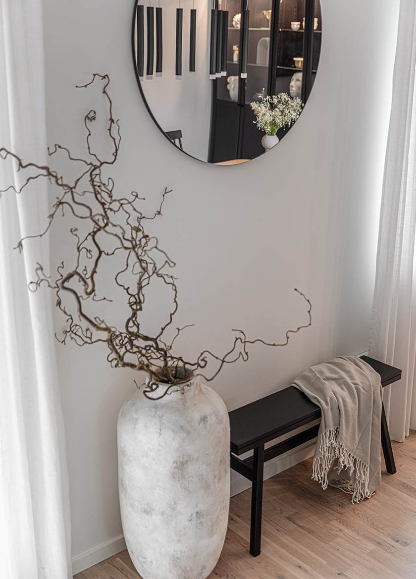 Stor grå vas med gren från trollhassel under rund spegel i vardagsrum
