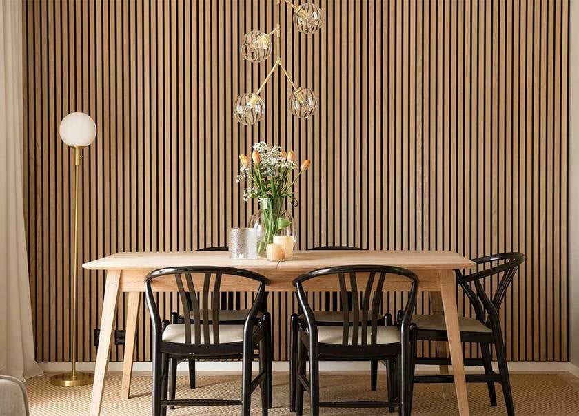 Akustikpanel i trä på vägg i vardagsrum. Framför står ett matsalsbord i ljust trä med svarta karmstolar runtom, i design som påminner om Y-stolen. 
