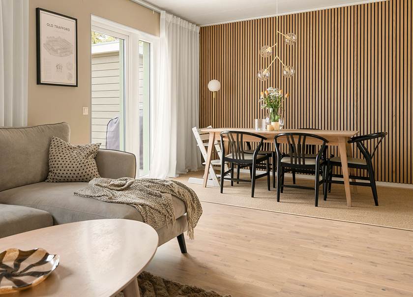 Akustikpanel i trä på vägg i vardagsrum med matsalsbord i ljust trä och ljusgrå soffa.