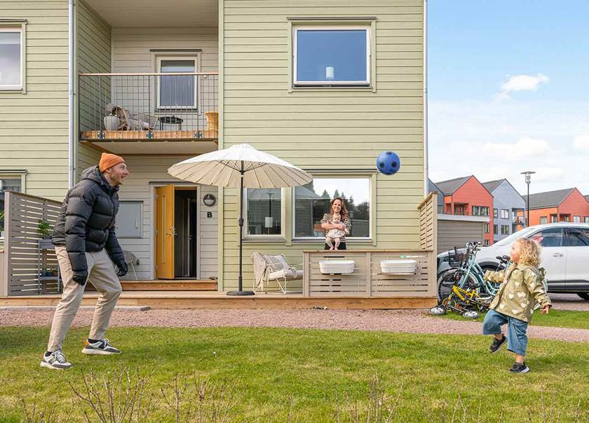 Vy av parhusets framsida med sin balkong och uteplats. Framför spelar en man och liten flicka fotboll.