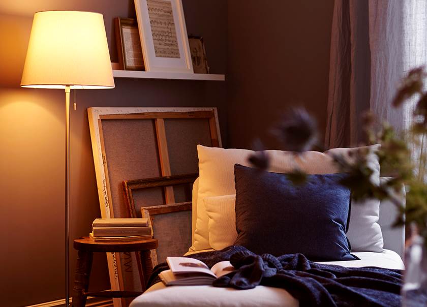 Tänd golvlampa med vit skärm, vid divan och tavlor vända bak och fram i ett hörn av  ett rum med gråa väggar.