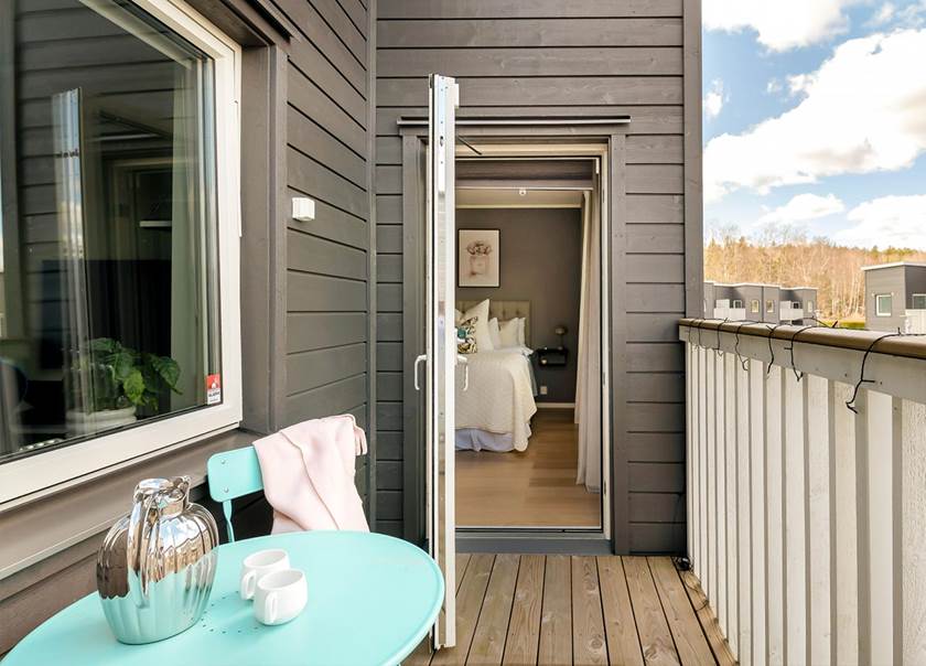 Vy på en balkong mot en balkongdörr i ett hus med grå träpanel på balkongen står ett turkos bord med silverkanna och vita koppar på.