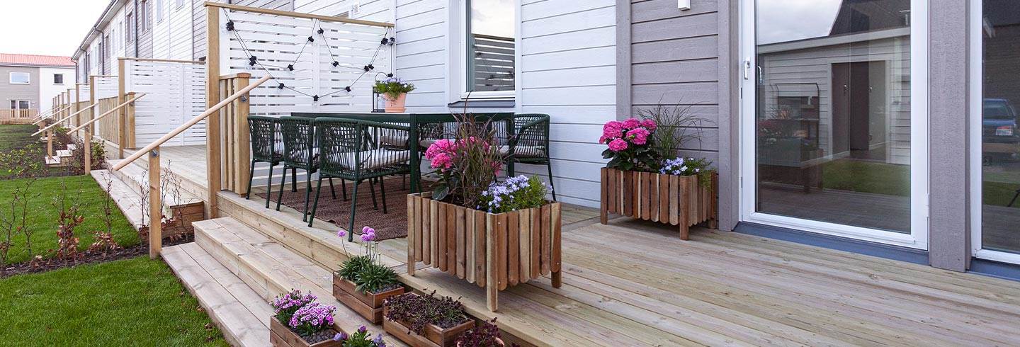 Lång terrass med grönt matbord och stolar. En bit ifrån står odlingslådor med gröna och rosa blommor.