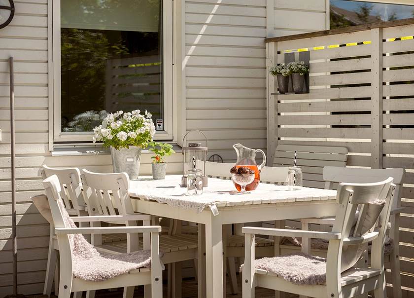 Vitt matbord och vita stolar på terrass. På bordet ligger en vit spetsduk. En karaff med röd saft står på bordet.