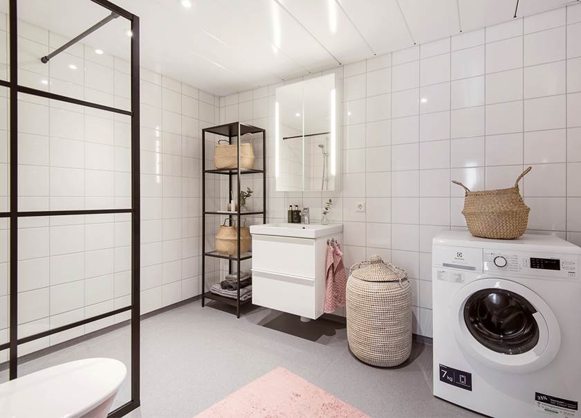 Vitt badrum med duschvägg i svart ram och klarglas, tvättmaskin och korgar i naturmaterial.