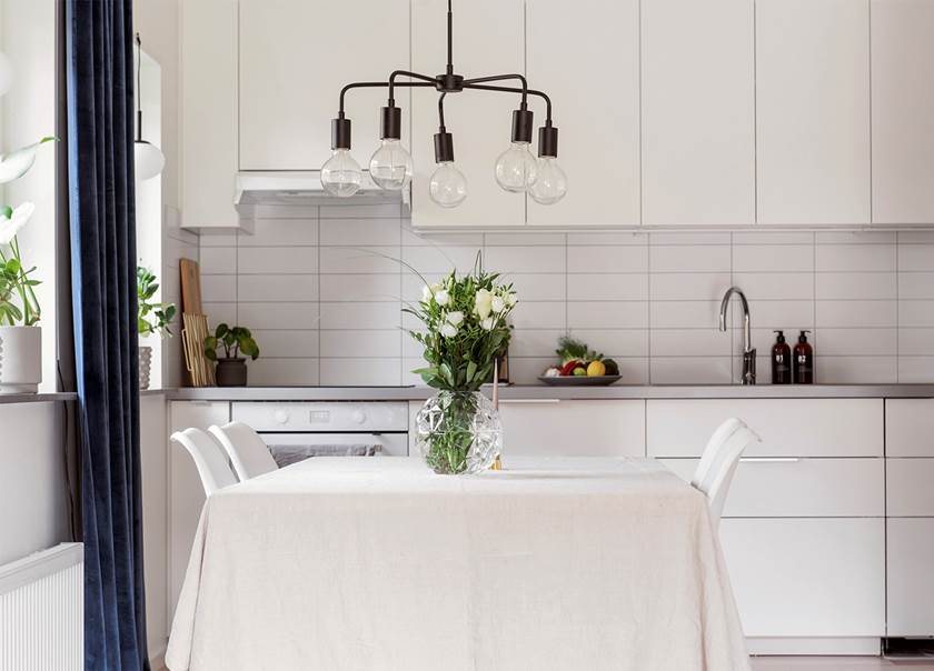 Svart femarmad taklampa ovan vitt matbord med vit duk och vita stolar i kök vid fönster med långa, blå sammetsgardiner.