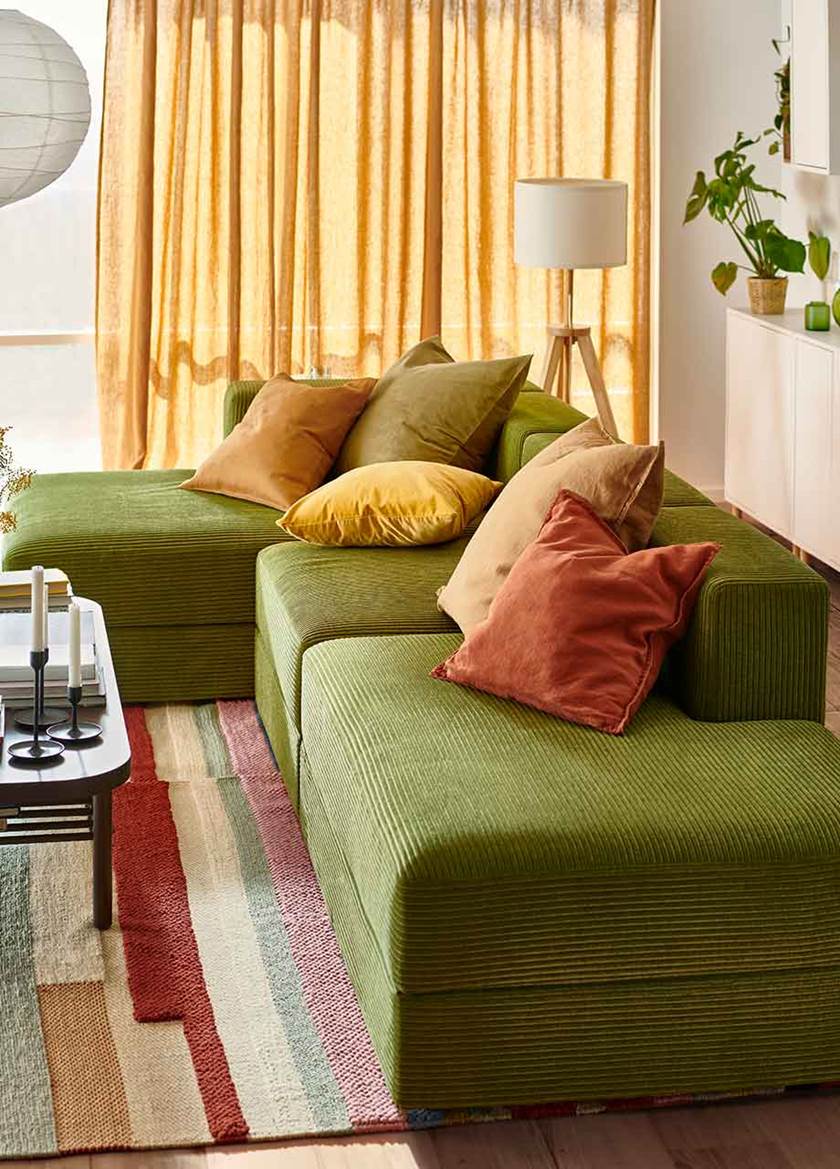 Grön soffa JÄTTEBO med kuddar i orange och gult på en randig matta i mörkorange, gult, rosa. Soffbord i mörkbrunt trä. Gardiner i gulorange ton.