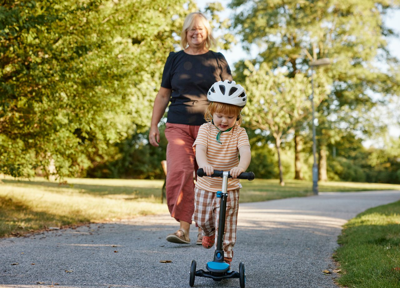 Foto på ett barn som åker sparkcykel med mormor eller farmor som promenerar bakom.bakom
