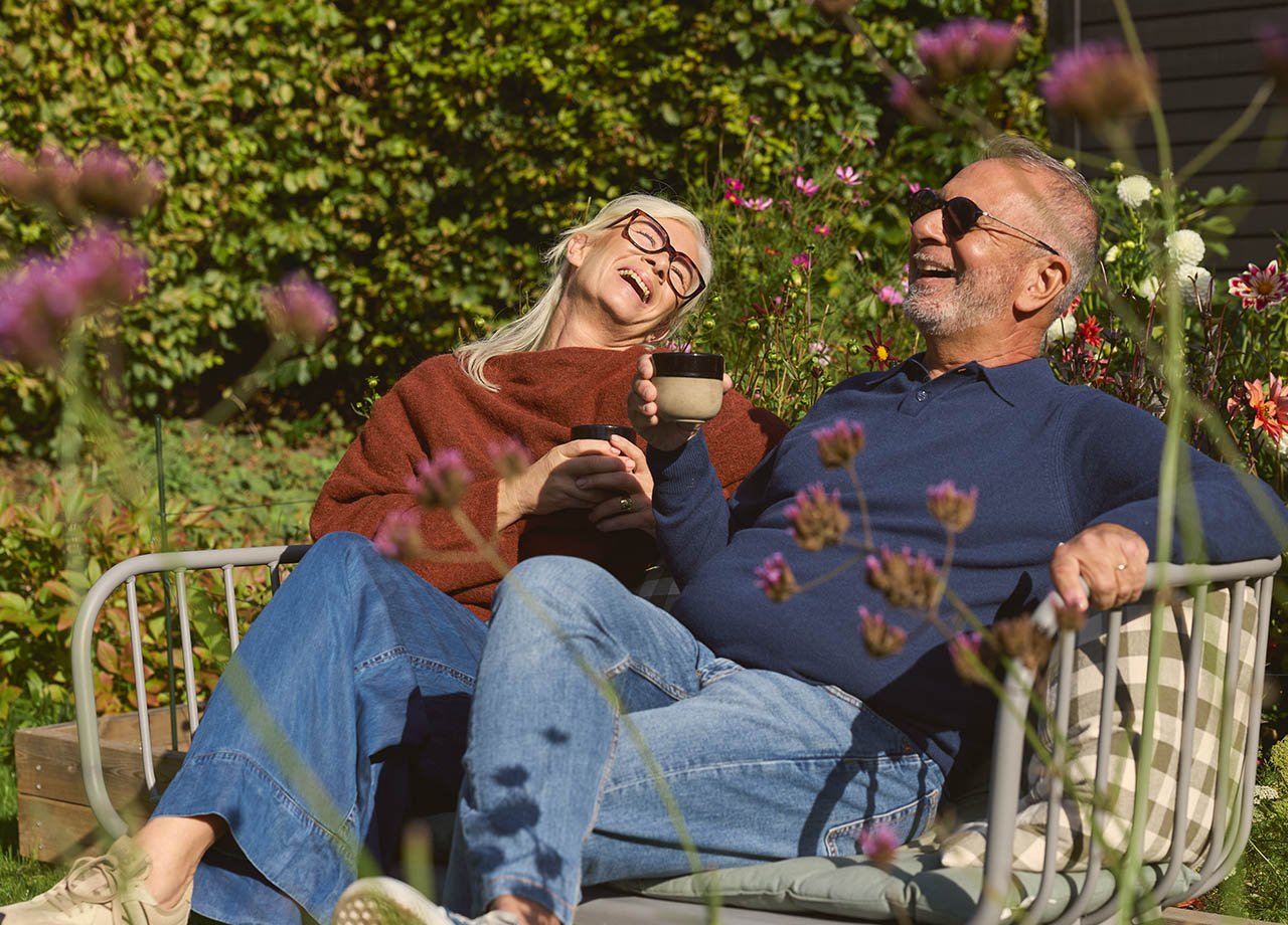 Skrattande äldre par sitter på bänk i trädgård.