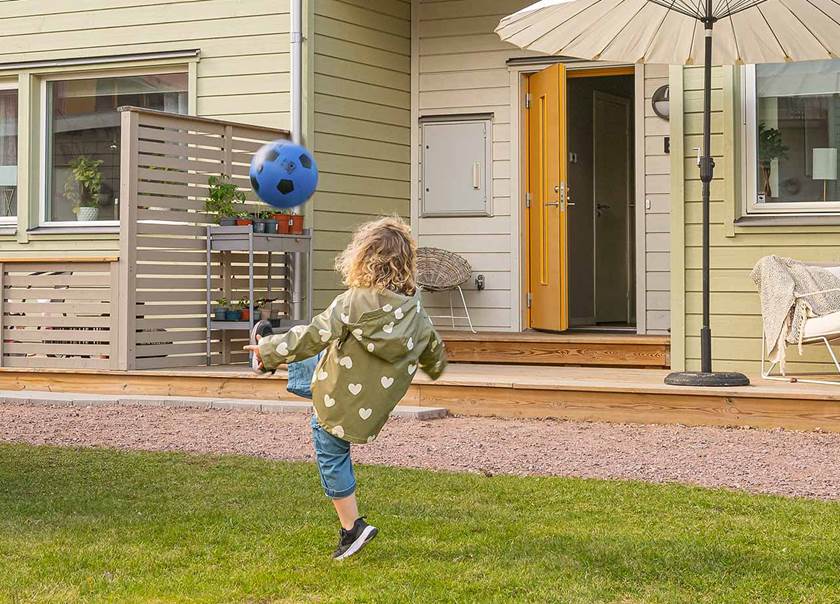 Barn sparkar boll på grön gräsplätt framför entré till radhuslänga