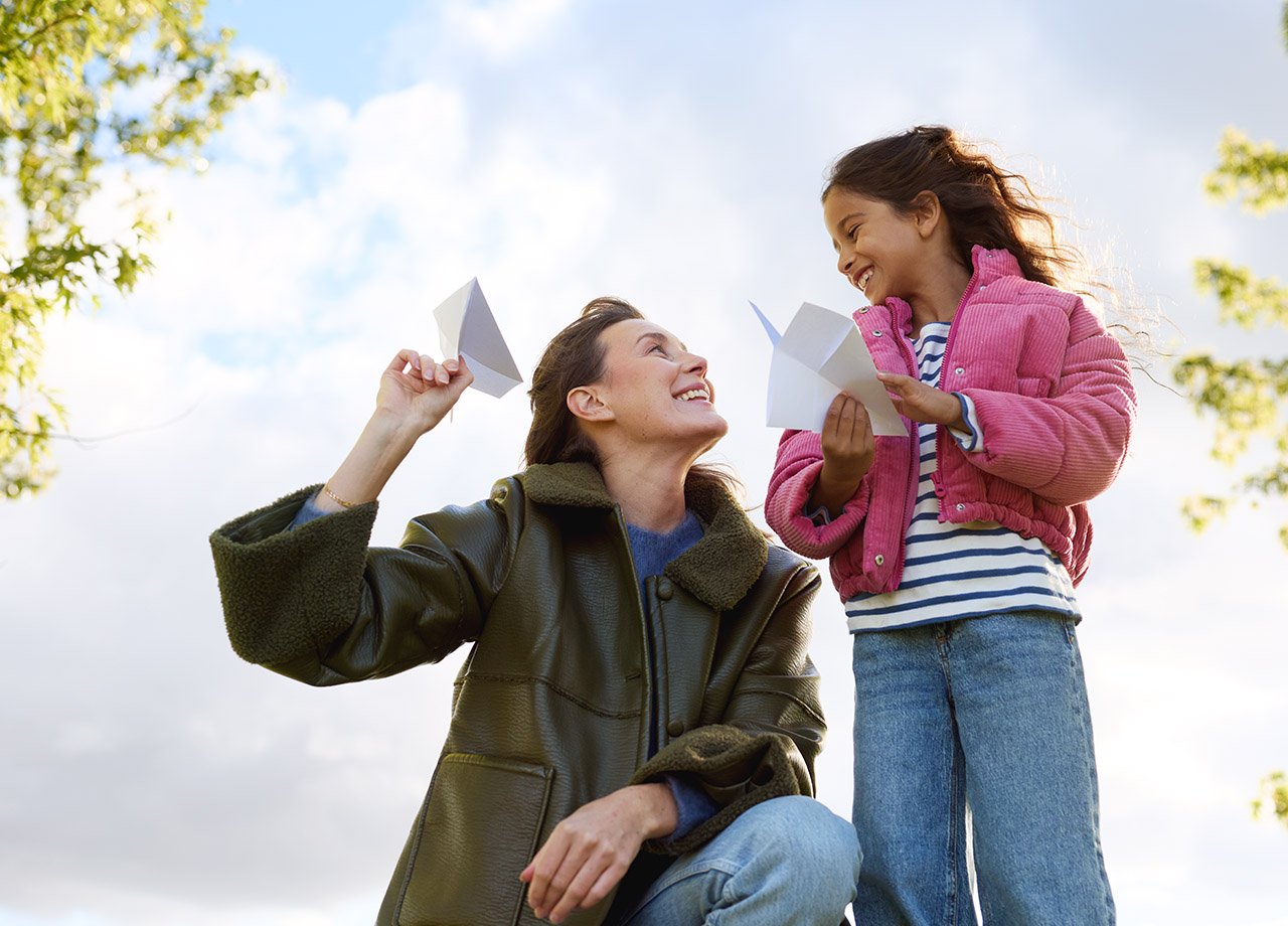 Mamma och dotter leker utomhus med pappersflygplan.