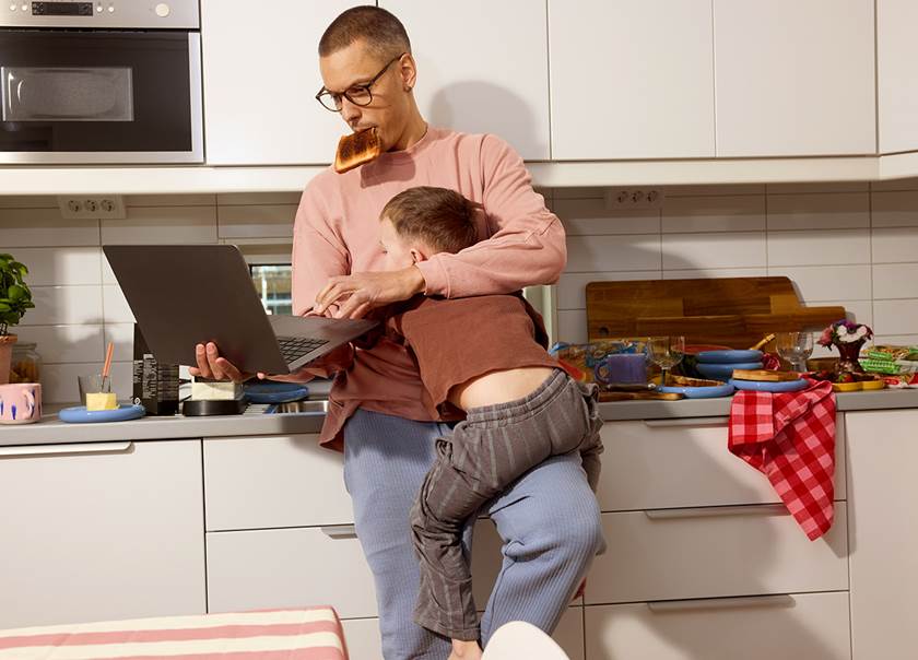 Pappa med son i famnen hållandes i laptop i stökigt kök.