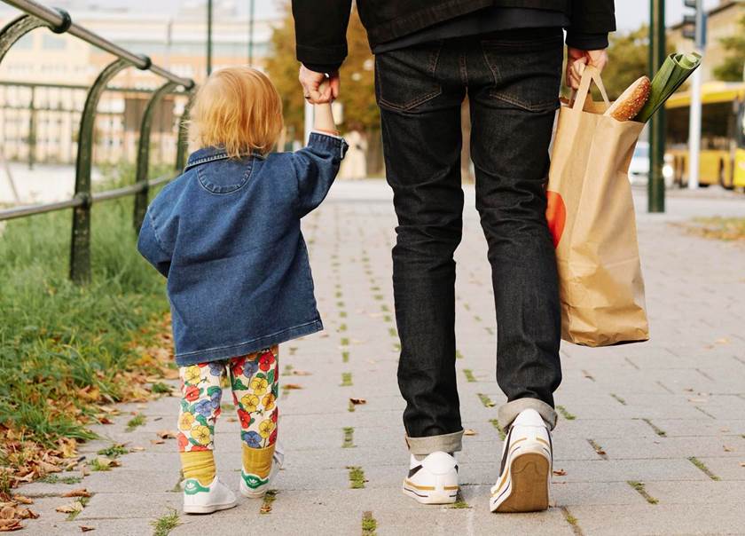Pappa och litet barn håller varandra i handen på promenad. Pappan håller matkasse i andra handen