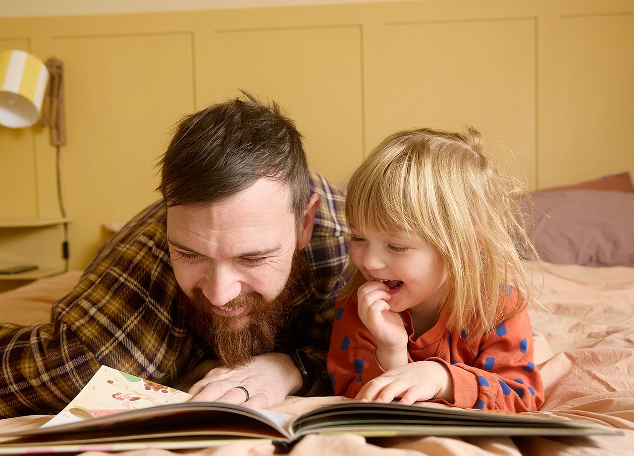 Pappa och dotter läser bok liggande på magen i en säng.