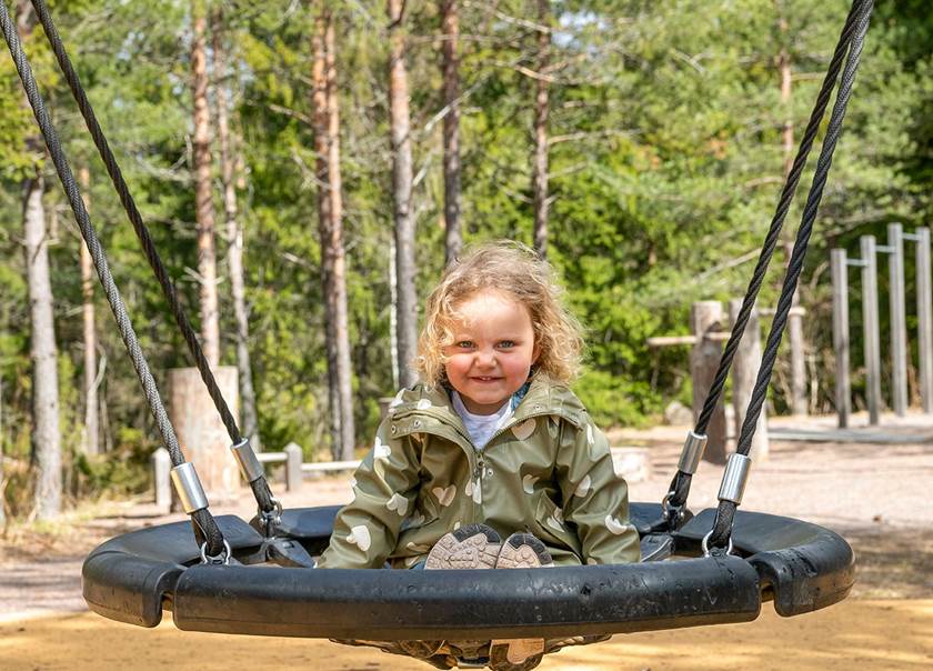 Flicka gungar på kompisgunga på lekplats med skog runt om